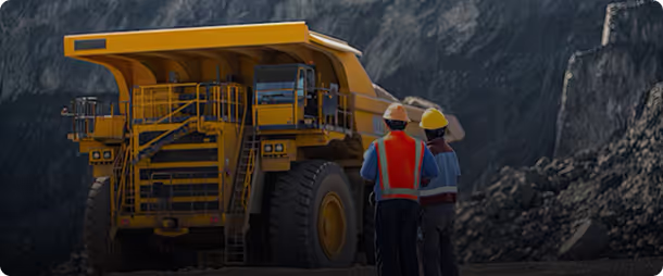 Two mine workers in safety gear standing near a large yellow mining truck at a mining site.
