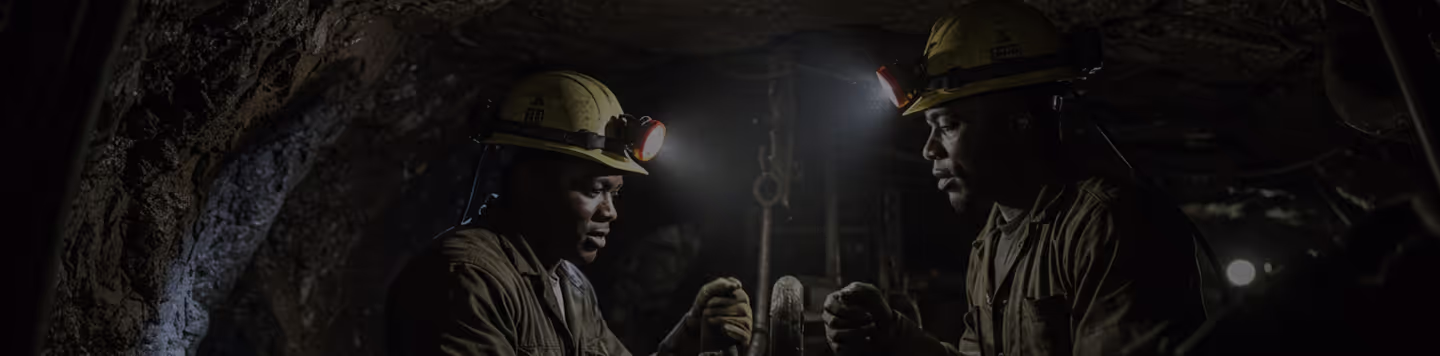 Two individuals wearing hard hats with lights work in a dimly lit underground mine.